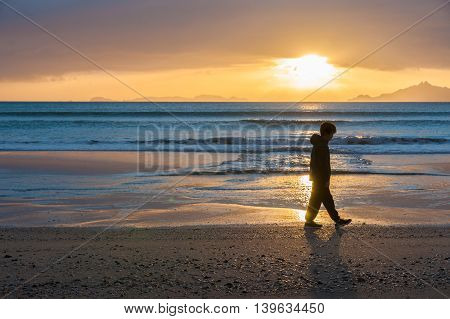 Sunrises glistening across water onto beach as a lone boy walks silhouetted at waters edge and offshore islands seen from Waipu Cove New Zealand.