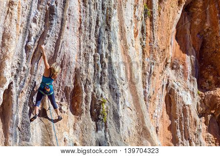 Blond Female Climber ascending Vertical Orange Rocky Wall unusual shape of Mountain Range on background Outdoor Sunshine