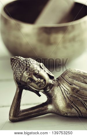 closeup of a representation of the buddha laying down, on a pale green rustic background, and a singing bowl with its mallet in the background