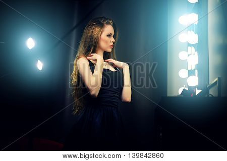 Beautiful sexy young caucasian woman in black dress looking into makeup mirror at herself. Backstage dramatic shot.