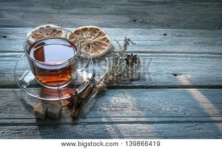 Transparent cup of black tea and dry twig herb dried oranges cinnamon sticks sugar and anise star on wooden background with copy space. Toned