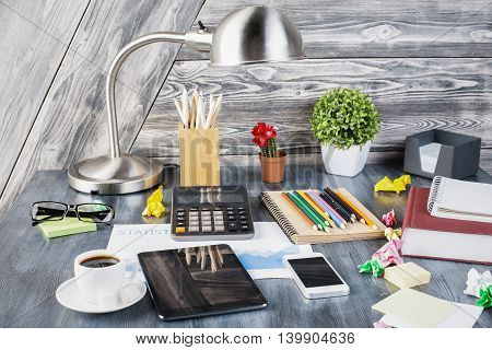 Closeup of creative messy desktop with blank tablet smartphone calculator table lamp decorative plants glasses various stationery items and other objects on wooden plank background. Mock up