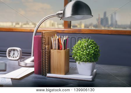 Closeup of table lamp notepads book pencil holder clock and plant on dark wooden office desktop. Window with city view in the background
