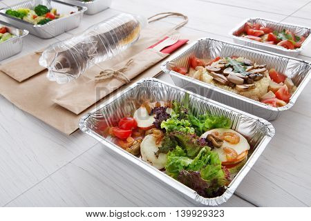 Healthy food and diet concept. Take away of fitness meal. Weight loss nutrition in foil boxes. Vegetables, lettuce and mozarella cheese with shrimps, paper bag and water bottle at white wood