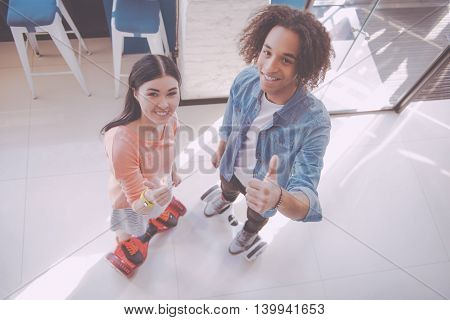 Good. Happy and cheerful young people riding hoverboards while being indoors in a cafe, posing and showing thumbs up