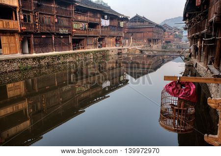 Zhaoxing Dong Village Guizhou Province China - April 9 2010: Wooden houses of farmers reflected in the water of the rural river cage with a bird hanging outdoors.