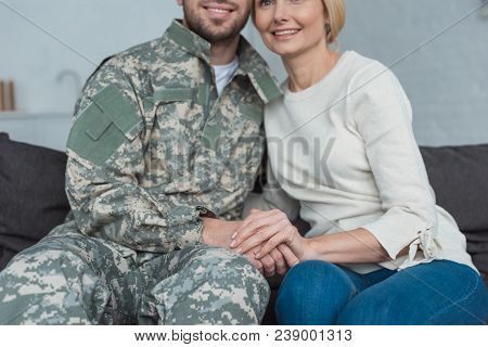 Partial View Of Smiling Mother And Grown Son In Military Uniform Holding Hands On Sofa At Home