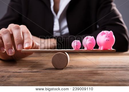 Businessperson's Finger Balancing Piggybanks On Seesaw