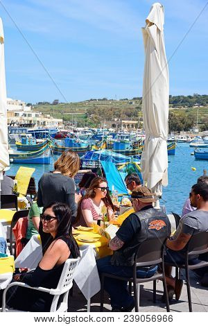 Marsaxlokk, Malta - April 1, 2017 - Tourists Relaxing At A Pavement Cafes Along The Waterfront With 