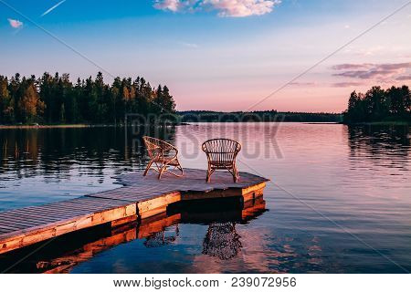 Two Wooden Chairs On A Wood Pier Overlooking A Lake At Sunset