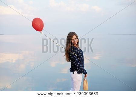 Portrait Of Young Woman With Red Air Balloon And Present Bag Near The Calm Sea Or Lake. Clouds Are R