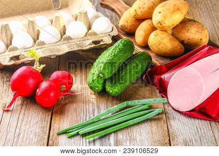 Ingredients For Okroshki - Sausage, Eggs, Radishes, Onions, Potatoes, Cucumbers On An Old Wooden Tab