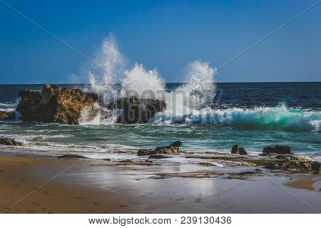Beautiful Beachfront View Of Waves Crashing Into Rock Formations On A Sunny Day With Stunning Blue W