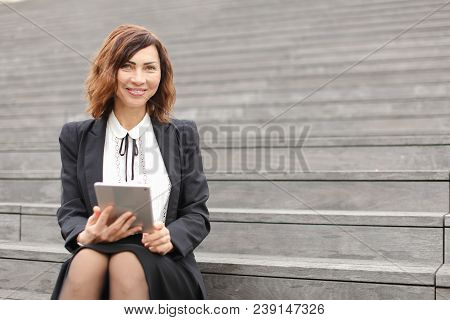 Female Smiling Secretary Sitting On Stairs With Tablet. Concept Of Working In Open Ait And Modern Te
