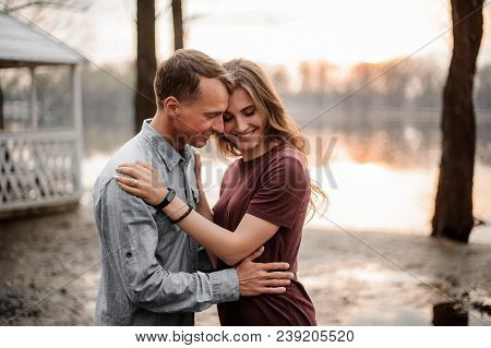 Couple In Love Holding Each Other Close On The Blurred Background Of Lake Sunset, Trees And Arbours 