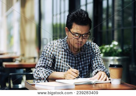 Vietnamese University Student Sitting In Outdoor Cafe And Writing In Texbook