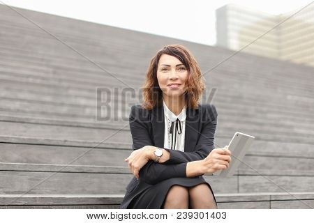 Caucasian Secretary Sitting On Stairs With Tablet. Concept Of Working In Open Ait And Modern Technol