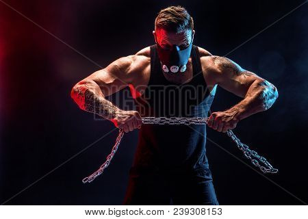 Portrait Of Muscular Sportsman In Training Mask Tearing Metal Chain. Black Background