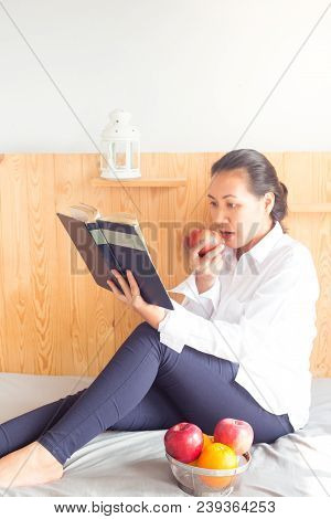 Asian Young Woman Reading Book And Eating Red Apple On Bed