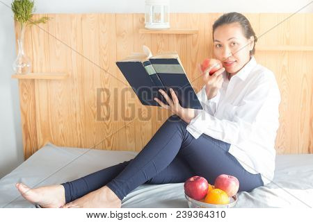 Asian Young Woman Reading Book And Eating Red Apple On Bed