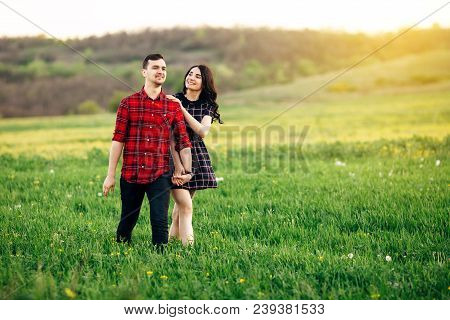Beautiful Couple Smoling And Holding Their Hands Walking At The Green Blooming Field At Sunset. Outd