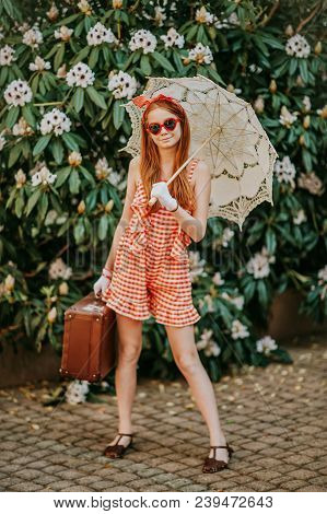 Outdoor Portrait Of Funny Little Redheaded Girl Wearing Red Gingham Playsuit, Holding White Lace Par