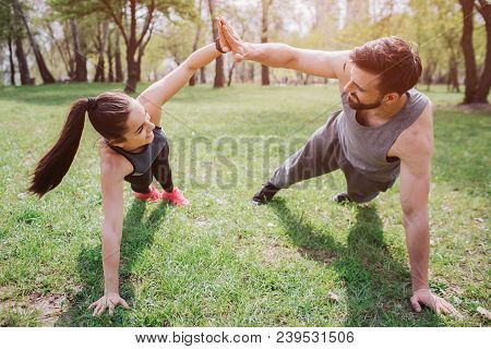 Slim And Well-built People Are Exercising On Grass In Park. They Are Staying In Plank Position And G