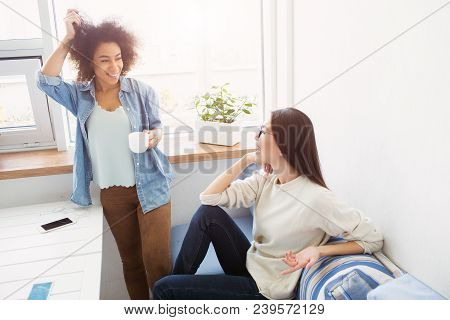 Two Students Are Talking To Each Other. Afro American Girl Is Standing Near Window And Touching Her 