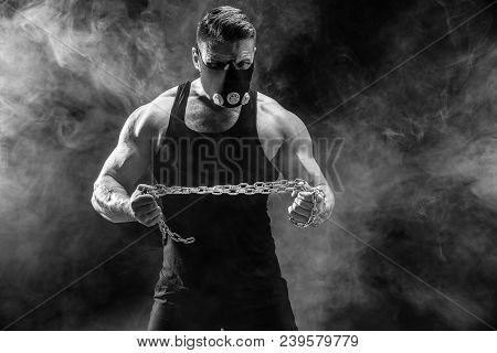 Portrait Of Muscular Sportsman In Training Mask Tearing Metal Chain.black Background With Smoke