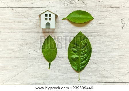 Miniature White Toy Model House With Green Leaves On Wooden Backgdrop. Eco Village, Abstract Environ