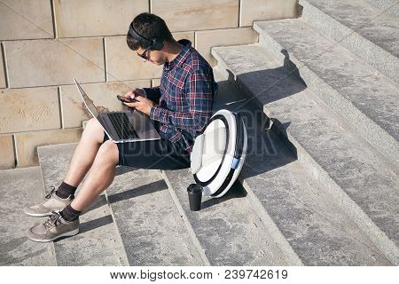A Man Is Sitting On The Stairs In The Street. Holds A Laptop And A Phone. Next To Him On The Stairs 