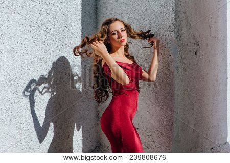 Beautiful Young Woman In A Long Red Evening Gown With A Train Standing Next To The Old Wall