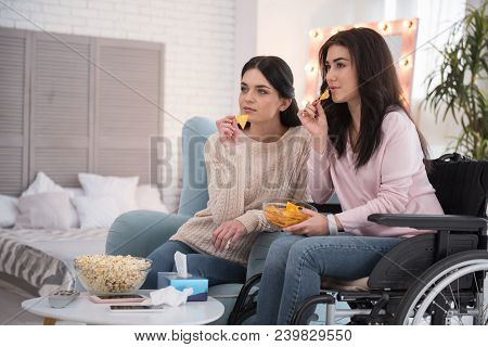 Sisters Friendship. Attentive Sister And Immobile Woman Eating Cheeps And Regarding Film