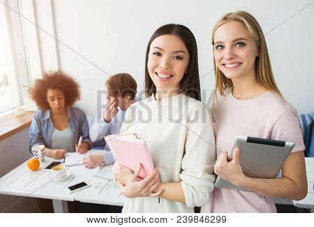 A Picture Of Two Beautiful Asian Girls Standing And Smiling. Brunette Holds A Pink Notebook While Th