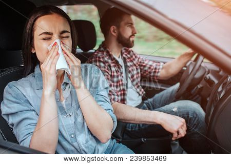 A Picture Of Sick Girl Riding In Car With Young Man. She Is Sneezing In Napkin While He Is Paying At