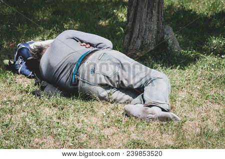 Old Barefoot Homeless Or Refugee Man Sleeping On The Grass In The Park Using His Travel Bag As Pillo