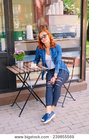Full Body Imgae Of Redhead Female Drinks Coffee At The Table In A Cafe On A Street.