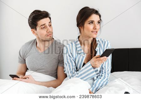 Portrait of a jelous young man looking at his girlfriends mobile phone while sitting together in bed at home