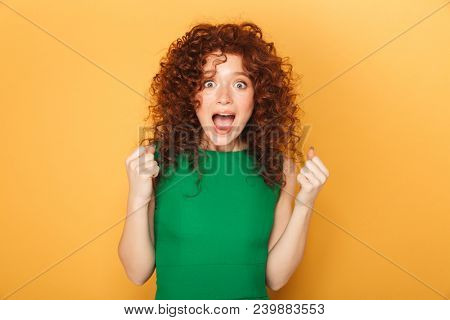 Portrait of a happy curly redhead woman celebtrating success isolated over yellow background