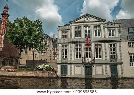 Bruges, Belgium - July 05, 2017. Wooded Gardens And Brick Buildings On The Canal Edge In A Sunny Day