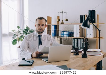 Portrait Of Lawyer With Documents Sitting At Workplace With Laptop In Office