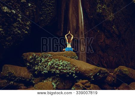 Woman Sitting On The Rock, Practicing Yoga. Young Woman Raising Arms With Namaste Mudra Near Waterfa