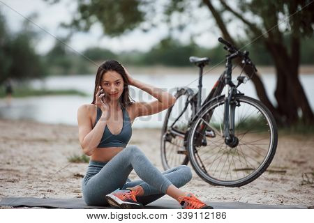 Having Conversation. Female Cyclist With Good Body Shape Sitting Near Her Bike On Beach At Daytime.