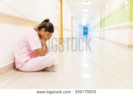 Stressed Young Nurse Covering Face While Sitting On Floor In Corridor At Hospital