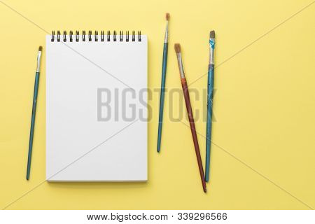 White Clean Sketchbook On Black Spring With Four Dirty Synthetic Brushes On Bright Yellow Paper Back
