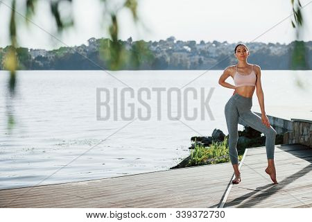 Full Length Photo. Young Woman With Slim Type Of Body Does Exercises Against Lake.