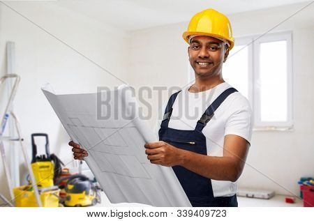profession, building and repair concept - happy smiling indian builder or repairman in helmet with blueprint over empty room with construction equipment background