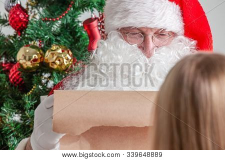 Santa Claus With With List Or Nice Or Naugthy List Paper With Small Boy Near Christmas Tree