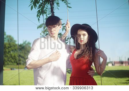 Beautiful Fashionable Couple On The Beach Near The River. Fashionable Hipsters. Girl In A Red Dress 