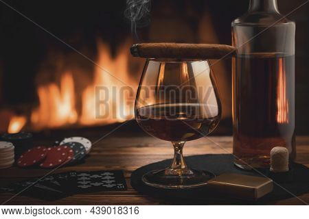 Glass Of Cognac, Cigar, Bottle, Cards Game And Chips On The Table Near The Burning Fireplace. Rest, 